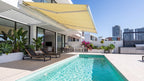 Cream folding-arm awning with white articulated arms above a poolside terrace; illustrative product view