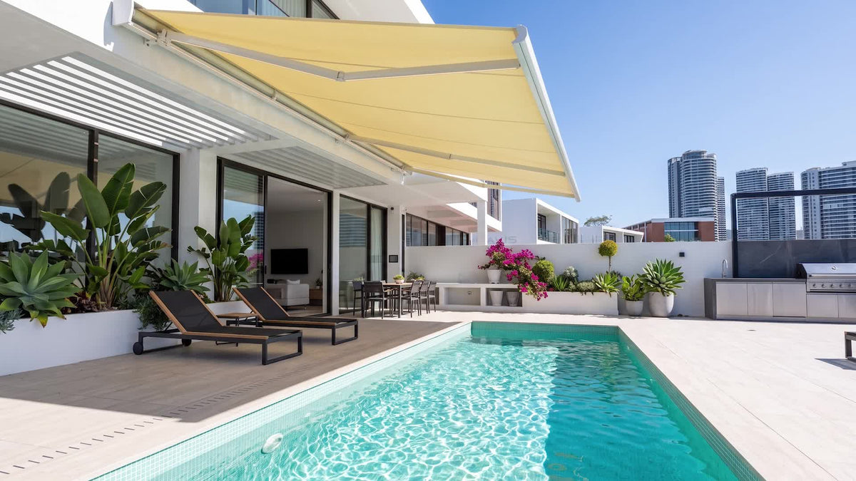 Cream folding-arm awning with white articulated arms above a poolside terrace; illustrative product view